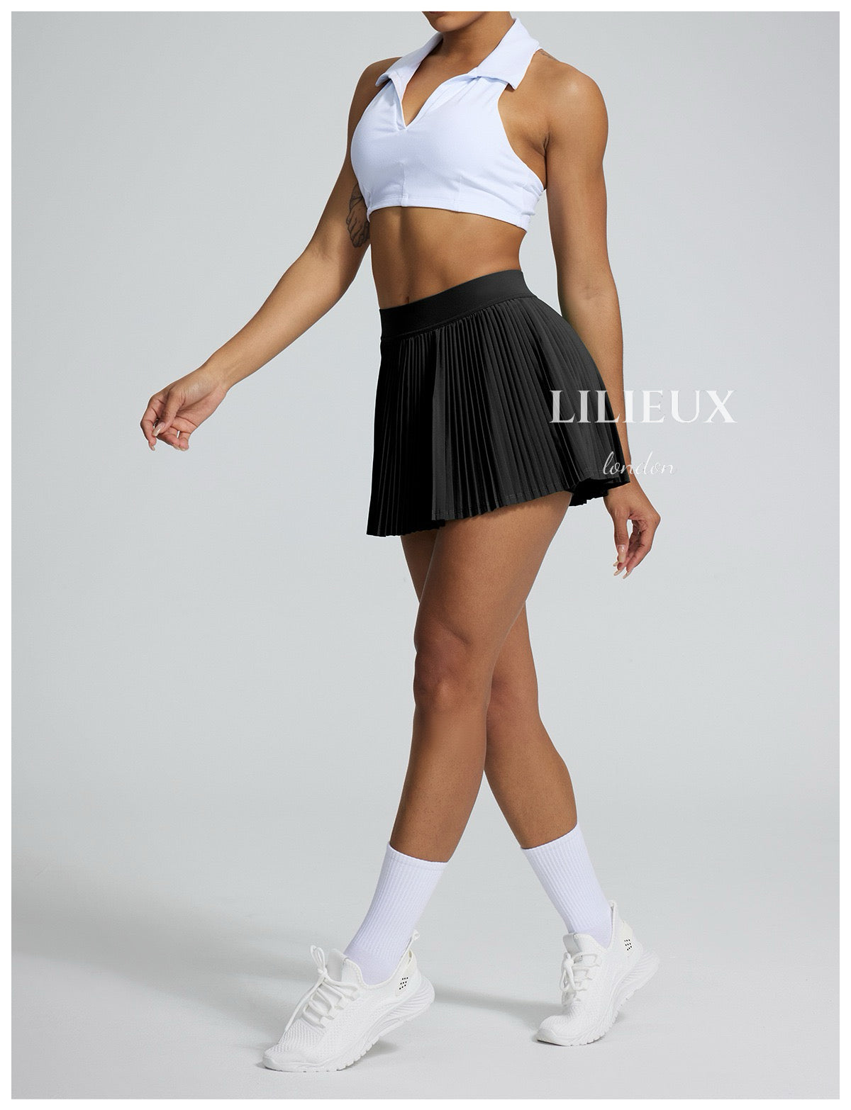 Female tennis player in a white sports bra and black pleated skirt, paired with white sneakers and socks, showcasing a sleek and sporty look.