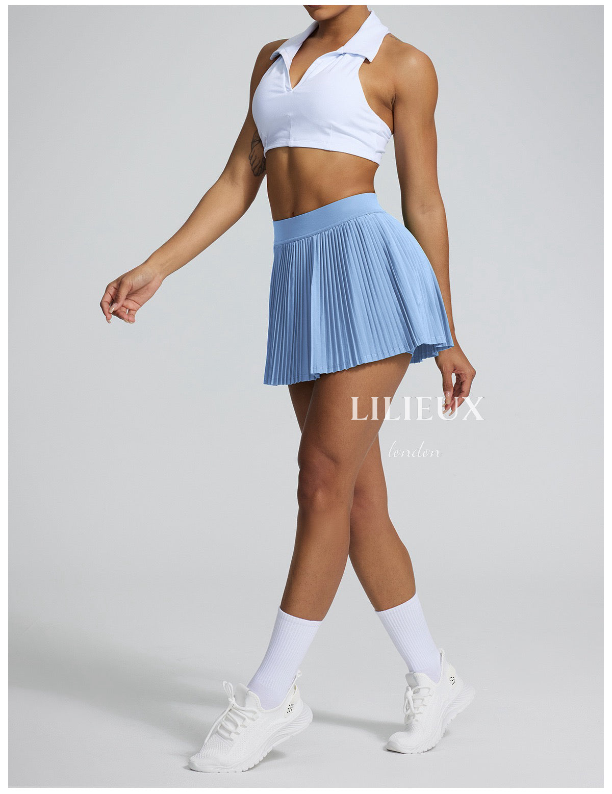 Female tennis player in a blue pleated skirt and white sports bra, paired with white sneakers and socks, posing in a sporty and stylish look.