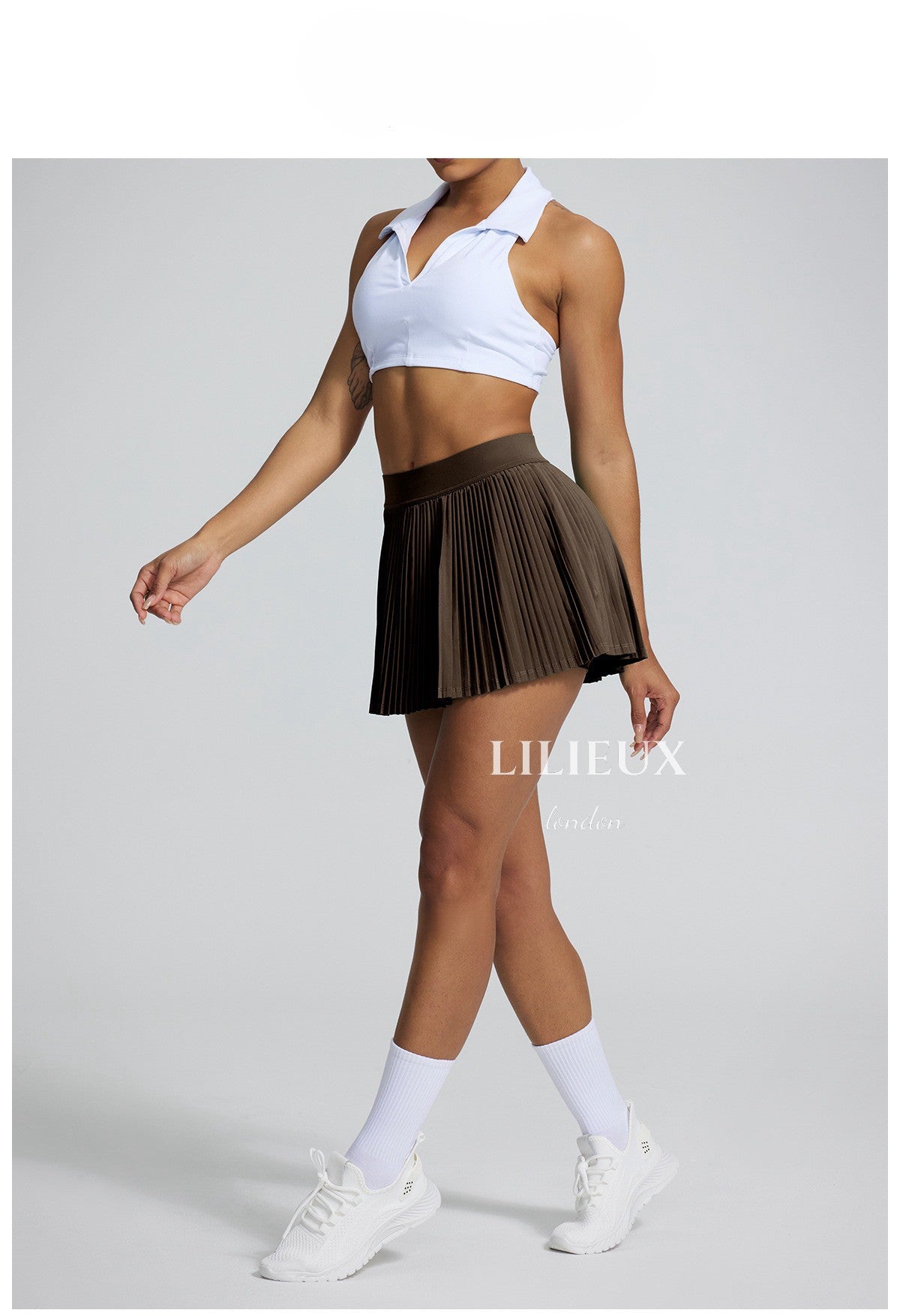 Female tennis player wearing a white sports bra and brown pleated skirt, paired with white sneakers and socks, showing a stylish athletic look.