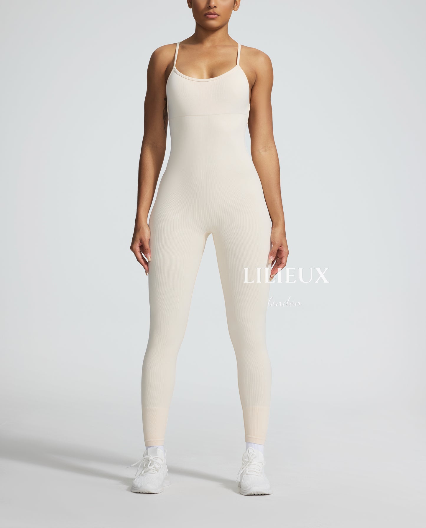 Female model wearing a cream-colored textured sports romper with thin straps, paired with white sneakers, showcasing a sleek and athletic look.