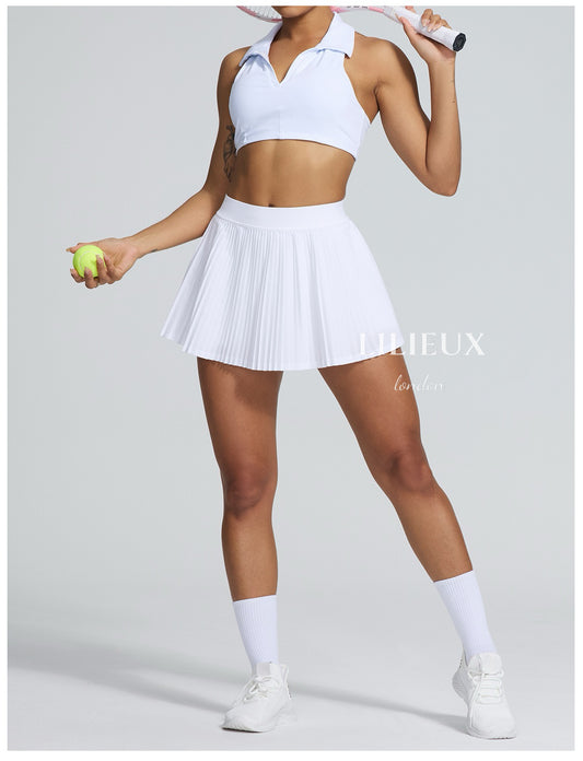Female tennis player wearing a white pleated skirt, white sports bra, and white sneakers, holding a tennis racket and ball in a studio setting.
