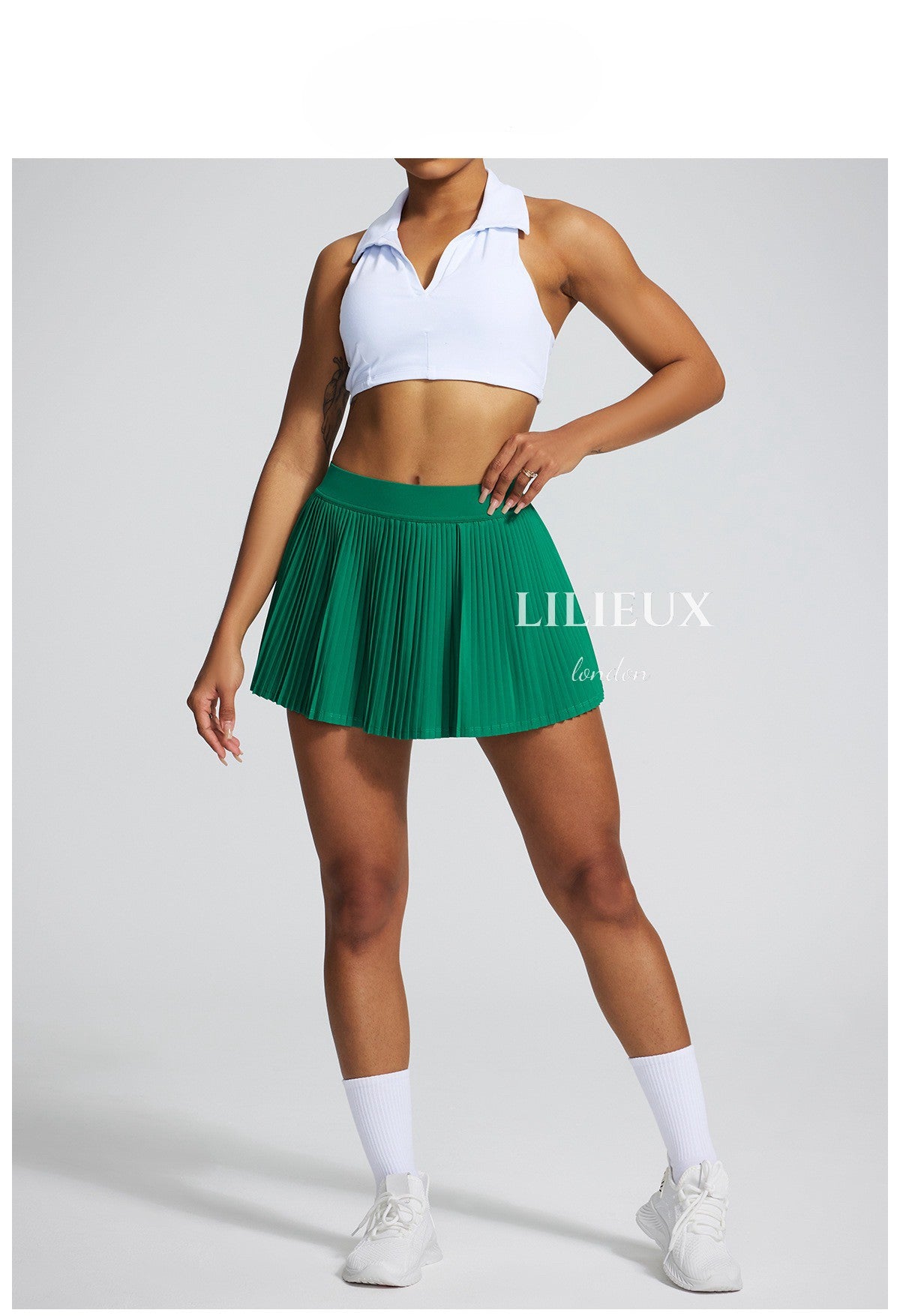 Model wearing a white sports bra and green pleated tennis skirt with white sneakers and socks, highlighting an athletic and fashionable ensemble.