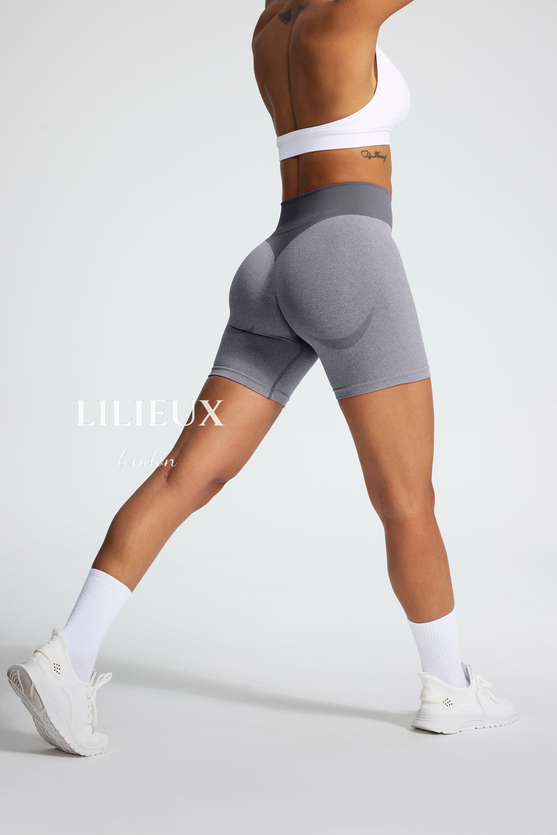 Female model wearing grey seamless butt-lifting biker shorts with a high-waist ribbed design, paired with a white sports bra and sneakers, showcasing a sculpted fit for activewear.