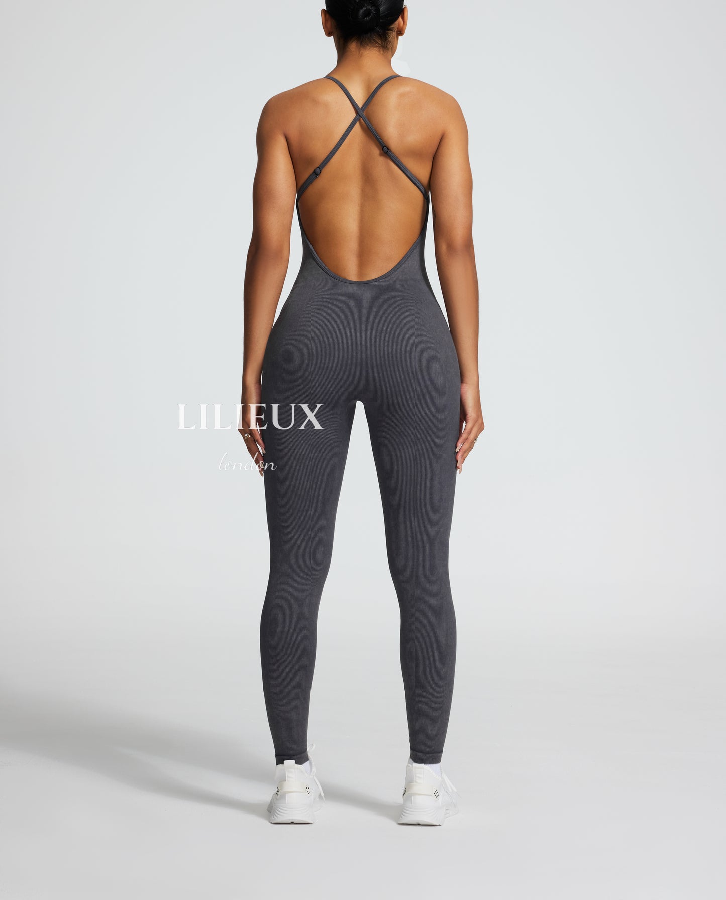 Back view of a female model wearing a grey sports romper with a crisscross back design and textured pattern, paired with white sneakers, highlighting a trendy athletic style.