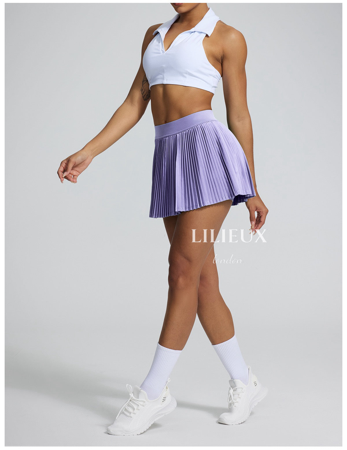 Model in a white sports bra and lavender pleated tennis skirt, with white sneakers and socks, showcasing a sporty and fashionable ensemble.