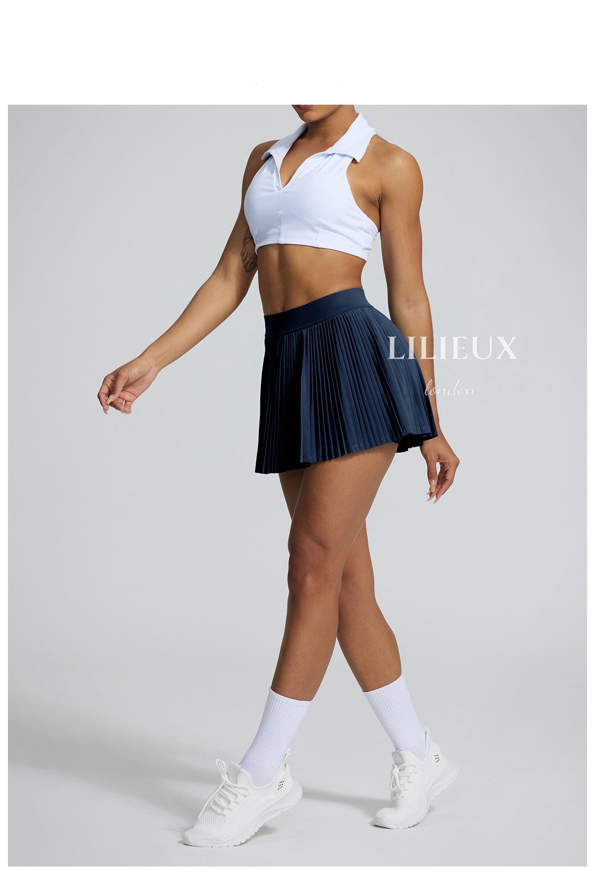 Female tennis player wearing a white sports bra and navy pleated skirt, paired with white sneakers and socks, showcasing a classic and sporty look.