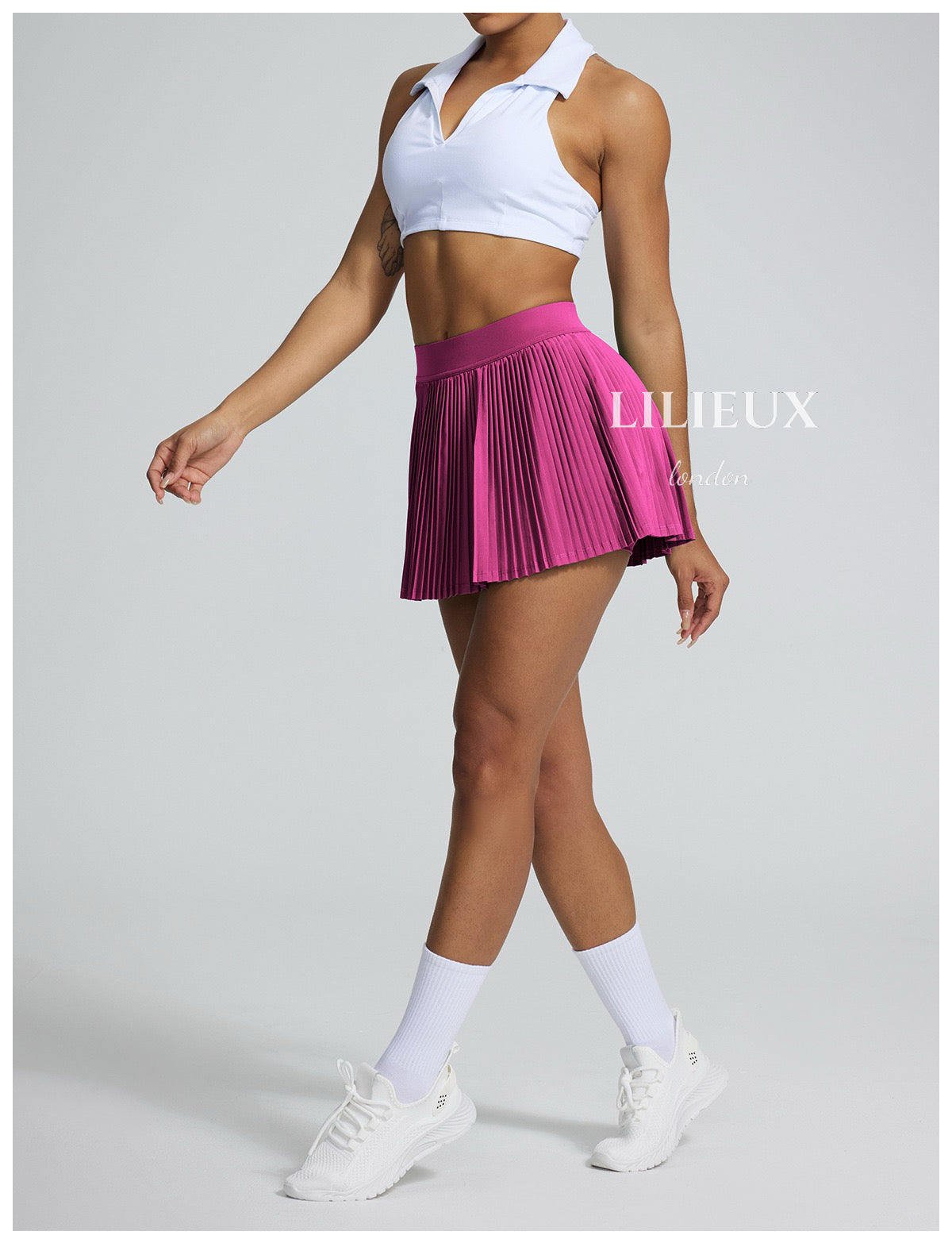 Female tennis player wearing a white sports bra and pink pleated skirt, paired with white sneakers and socks, presenting a vibrant and sporty look.