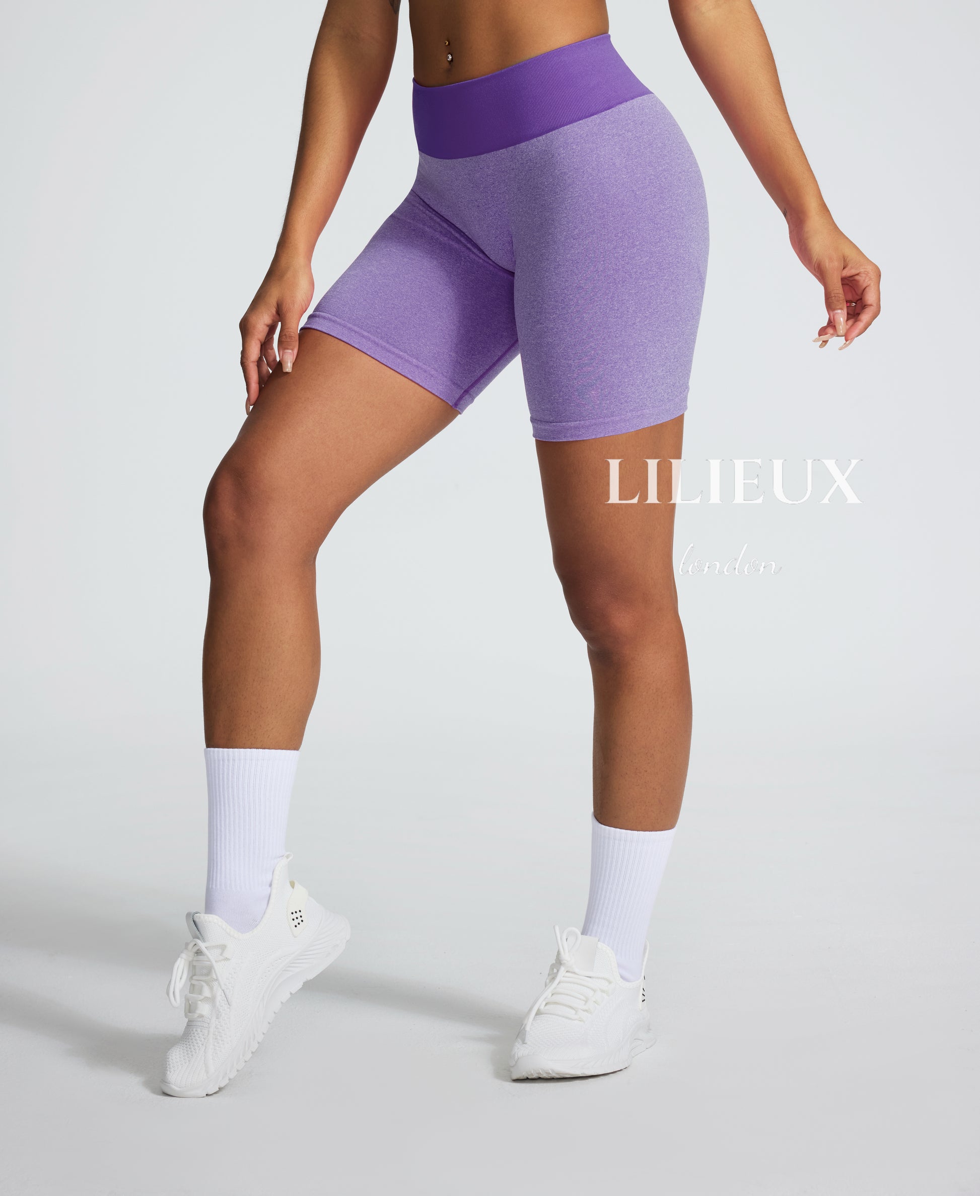 Female model wearing high-waisted purple seamless biker shorts with a ribbed waistband, paired with white sneakers and socks, designed for gym and activewear fashion.
